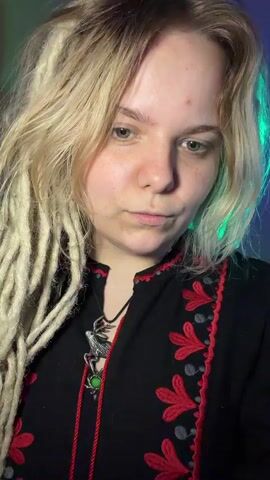 The video displays a close-up shot of a young woman with blonde dreadlocks and fair skin. She is wearing a black garment adorned with red floral embroidery and a silver pendant featuring a dragon and a green stone. The lighting in the scene is dramatic, with a green light illuminating the right side of her face and hair, contrasting with a darker background.

The woman is actively engaged in playing a musical instrument, likely a jaw harp or jew's harp. She holds the instrument to her mouth with her left hand, while her right hand plucks a small metal tongue protruding from the instrument. Her lips are slightly parted, and her eyes are partially closed, suggesting concentration on the music she is creating. The movement of her hands and the instrument is the primary focus of the action.

The setting appears to be indoors, with the backdrop dimly lit and tinged with the green light. There are no overt clues about the specific location or time of day beyond the indoor setting and artificial lighting. The mood is focused and artistic, centered around the performance of the musical instrument. No visible text is present in the video.