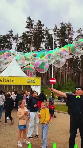 An outdoor scene features a man in a black hat, sunglasses, black shirt, and green bow tie, holding a long stick with a bubble wand, creating numerous large, iridescent soap bubbles that float upwards against a cloudy sky. He is positioned in the right foreground on a paved surface. In the background, a dense line of tall pine trees is visible. To the left, a white tent with a yellow "IKEA" logo is partially visible. A red and white "no entry" sign is mounted on a pole near the center of the frame. Several adults and children are gathered, observing the bubbles and the man; some children are looking up and reaching towards the bubbles. The ground in the mid-ground appears to be grass, with some small colorful tables or stools visible on the right.