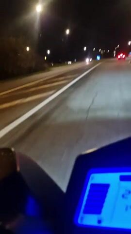 A first-person perspective video shows a motorcycle or scooter riding on an asphalt road at night. The blue digital speedometer is visible in the foreground. Streetlights line the road, and other cars are visible in the distance, their headlights and taillights illuminating the scene. The road has white lane markings. The surrounding environment appears to be a suburban or semi-urban area with some vegetation visible alongside the road. The vehicle appears to be traveling at a moderate speed.