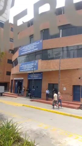 The video shows the front of a brick building identified as the "DELEGACION ESTADAL LA GUAIRA" and "DELEGACION MUNICIPAL LA GUAIRA." Several people are standing and walking around the entrance, including a couple walking together across the sidewalk.  A motorcycle and a small white truck are parked nearby. Orange cones are placed on the street.  Trees and other greenery are visible in the foreground.  A taxi is parked on the street at the edge of the frame.  There are several signs with text on the building's facade.