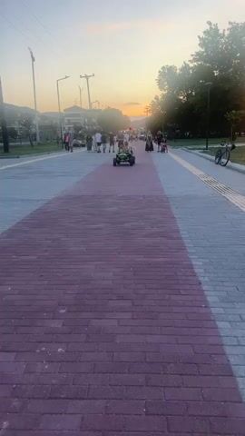 The video captures an outdoor scene with a paved walkway in the foreground divided by a red brick section. Several people are walking and standing on the walkway, while one person is operating a green go-kart with two children inside. The go-kart is moving forward on the red brick path. In the background, trees line the right side of the walkway, and buildings and utility poles are visible on the left. The sky is illuminated with the warm colors of sunset. A bicycle is parked on the right side of the walkway. The atmosphere appears to be casual and social, with people enjoying a leisurely evening outdoors.