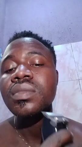 A man is shaving his face with an electric razor. He is a dark-skinned man with short, curly black hair. He has a thin mustache and a small goatee. He is shirtless and wearing a gold chain. He is looking down at the razor as he shaves. He is in a bathroom with a purple ceiling and a white tiled wall. The razor is black and silver.