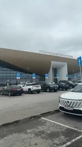 The video shows an exterior view of an airport with several cars parked in front of it. The airport building has a modern design with a large curved facade. The sky is overcast. There are no people visible in the video. The cars are parked in designated parking spots. The video is shot from a stationary position.