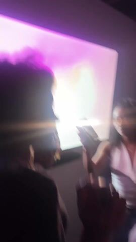 The video shows a dimly lit room with a pinkish hue, likely from artificial lighting. Several people are present, mostly visible in silhouette or with blurred features.  The focus is unsteady and shifts rapidly, giving a sense of movement and activity.  There is a white wall visible in the background.  Sounds suggest a party or event with music and enthusiastic singing or shouting.