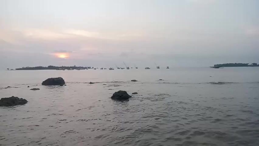 Here is a description of the video:

The video shows a calm sea with several boats on the horizon, their tall masts clearly visible against the faint, orange-tinted sky. In the foreground, there are scattered rocks partially submerged in the water, with small waves gently lapping around them. An island is visible in the distance to the right. The overall atmosphere is serene and peaceful.