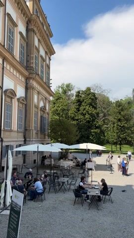 The video shows an outdoor cafe next to a large building in a park. The building is multi-storied with many windows and a tan and white facade. The cafe has several tables with chairs under white umbrellas. People are sitting at some of the tables, and others are walking around. A sign for the restaurant is visible. Beyond the cafe is a gravel path leading into the park, where there are many trees and grassy areas. People are walking and playing soccer on the grass. The sky is blue with some white clouds.