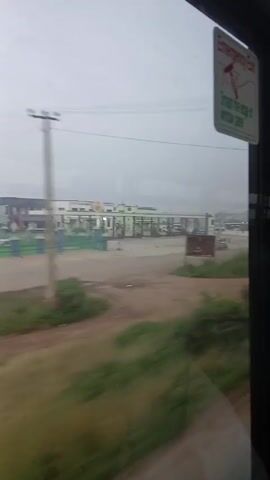 Okay, here is a concise description of the video:

The video is taken from inside a bus, looking out the window as it drives past a building on a hazy day.  The building appears to be a series of white and green structures, possibly toll booths or a service area of some kind.  Between the bus and the building is a patch of dirt and some green vegetation.  The bus's interior is visible, including an "Emergency Exit" sign and seats with colorful fabric. The camera moves slightly, providing a view of the bus's interior.