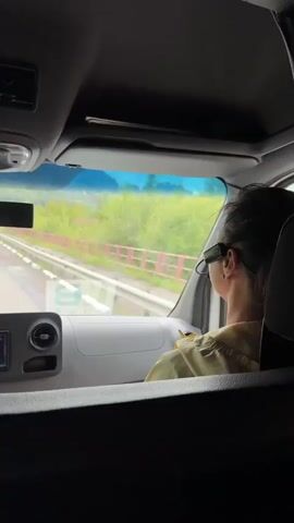 The video shows the interior of a vehicle, likely a van or bus, from the perspective of a passenger. The camera is positioned to capture the dashboard and the view through the windshield. A person, seen from behind, is in the driver's seat, wearing glasses and a light-colored jacket. Their legs, clad in blue jeans and sneakers, are visible. The dashboard features a multimedia system displaying the radio frequency 87.50, air vents, and storage compartments. Outside the windshield, a road stretches ahead, bordered by a guardrail and lush green trees and hills under a cloudy sky. The vehicle appears to be in motion, indicated by the blurred scenery. The text overlay in Russian translates to "We are moving along the left bank of Katun."