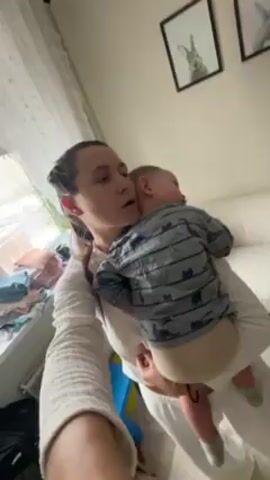 A woman with dark hair pulled back from her face holds a baby close to her chest. The baby is wearing a gray and white striped shirt with cat designs and a diaper. The woman is wearing a white shirt. They are in a room with white walls, a window with white curtains, a white couch, and two framed pictures of rabbits. The woman is looking down and appears to be shushing the baby. The camera angle is slightly upward, as if the person is taking a selfie video.