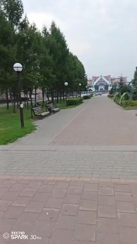 The video shows a paved walkway lined with trees on both sides. Benches are visible along the left side of the walkway. In the distance, a building with a distinctive architectural style, possibly a train station or a public building, can be seen. The sky is overcast. The timestamp indicates the video begins at 00:00 and ends at 00:04.