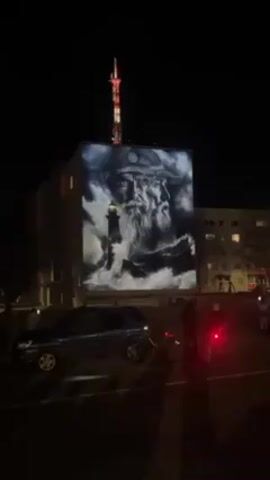 At night, a large animated image is projected onto the side of a tall building. The image depicts a bearded, uniformed man at sea, with a lighthouse shown at his side. There are clouds and waves in the image. At the top of the building, a tall red and white communications tower is visible. Several other multi-story buildings are visible in the background. In the foreground are several people standing on the street, as well as a dark SUV and a motorcycle. The motorcycle has a red light on its rear.