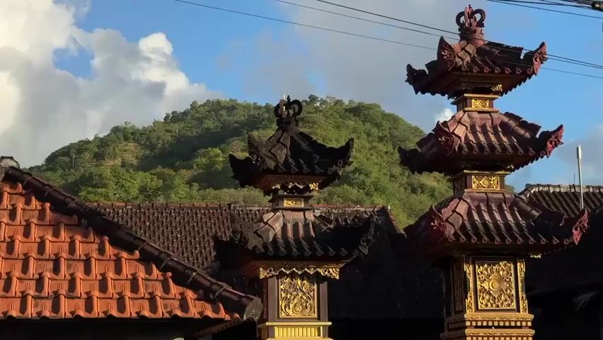 A close-up shot shows the ornate tops of two traditional Balinese structures against a backdrop of a green, tree-covered mountain. The structures are adorned with intricate dark red and gold carvings and are topped with tiered, pagoda-like roofs. To the left, a portion of a terracotta tiled roof is visible. The sky above is blue with scattered white clouds, and thin electrical wires are visible across the upper portion of the frame. The lighting suggests it is daytime, possibly late afternoon, with the sun casting a warm glow on the scene. There are no people or animals visible.