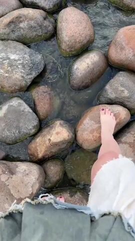 The video opens with a close-up shot of a person's feet and legs as they sit on the edge of a body of water. The feet are bare, and one foot has painted toenails. The legs are propped up, and the person is wearing a white garment that drapes over their legs. The water is shallow, with large, rounded rocks visible beneath the surface and along the shore.

The camera then pans upwards, showing the water flowing around more rocks. The scene transitions to a wider shot of a lake or large body of water with a rocky shoreline. The sky is filled with dramatic clouds, and the water is choppy, with small waves lapping at the shore. In the distance, a beach is visible with trees and buildings, and a lighthouse with red and white stripes stands prominently on the horizon. A faint rainbow can be seen in the sky to the left of the lighthouse. The overall impression is a view of a natural landscape at dusk or under a cloudy sky.