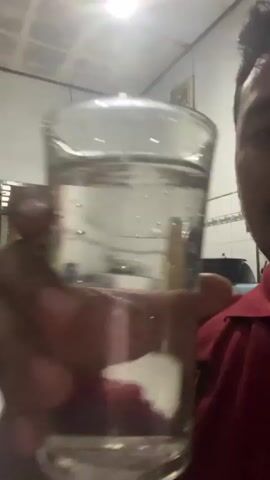 This is a close-up, first-person perspective video of a person drinking from a glass of clear liquid, likely water. The individual is wearing a red collared shirt with a black and red patch on the left chest area. The setting appears to be indoors, possibly a home, with white tiled walls and a white ceiling with visible light fixtures. In the background, a curtained doorway or window is visible, and further back, a tiled wall with some kitchen appliances or storage items can be seen, including what looks like a black pot or container. A framed picture hangs on the wall in the upper background. The lighting suggests it is daytime, but the interior setting makes it difficult to determine the exact time of day or weather conditions. The person's face is mostly obscured by the glass, but their hand and arm are clearly visible as they hold and drink from the glass. The overall mood is casual and everyday. There is no visible text in the video. The location context is Keudematangkuli, Indonesia.