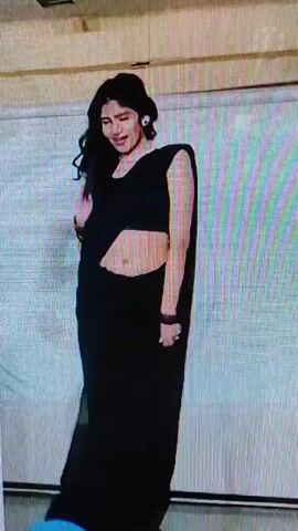 A full-body shot of a young South Asian woman dressed in a black sari and sequined blouse is seen dancing in what appears to be an indoor event space. The sari is draped to reveal her midriff, and she is wearing bracelets on her wrists. She smiles and gestures with her hands as she moves to the music, with her hair flowing freely. In the background, a white curtain or backdrop is visible, with a ceiling and lighting fixture at the top of the frame. Another person's shoulder and arm can be seen on the left side of the frame, suggesting the presence of other individuals in the vicinity. The video quality is somewhat grainy and low-resolution, with visible digital artifacts.