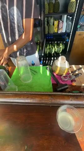 The video shows a person preparing a drink at an outdoor stand. The person, wearing a dark t-shirt with a circular design and white shorts, stands behind a wooden counter. In front of them is a green cloth with a plastic shaker, a small bottle with a dropper, and a halved coconut. Tools such as scissors and a hammer are also visible on the counter. In the background, shelves display pineapples, watermelons, and bottled drinks. A glass of a pale yellow liquid sits on the counter in the foreground. The person's hands are manipulating items on the counter. The scene appears to be a food or drink stall.