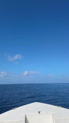 The video shows a view from the front of a white boat moving across a calm, blue ocean under a clear, blue sky with scattered clouds. The horizon is visible with a faint outline of land in the distance.