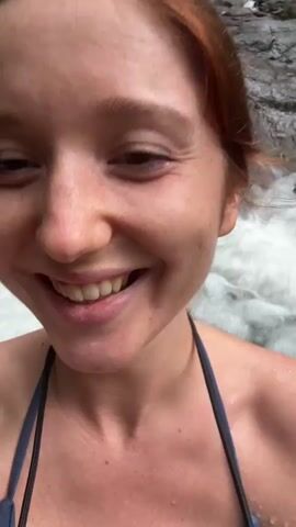 The video shows a woman with reddish-brown hair pulled back into a ponytail taking a selfie. She is smiling broadly, with water splashing around her. She wears a swimsuit with dark straps. Behind her, a waterfall cascades down a rocky cliff. Lush green vegetation surrounds the waterfall. Another person is visible on the rocks near the waterfall in the background.