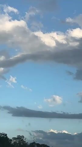 The video shows a cloudy sky. The sky is blue, with various gray and white clouds. A dark mass of trees is visible at the bottom of the frame. Electronic music can be heard in the background.