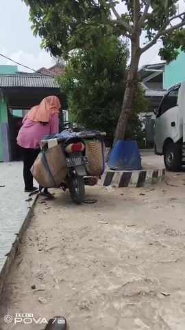 A person wearing a pink hijab and pink shirt with black pants is seen from behind, pushing a motorcycle. The motorcycle has two large woven baskets attached to the rear, one on each side, and a blue barrel is visible behind the motorcycle. The ground is dirt and sand. In the background, there is a tree, a building with green and blue paint, and a white truck. The sky is cloudy.