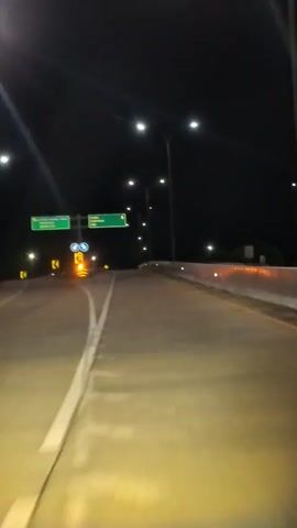 The video shows a first-person perspective from a vehicle driving on a well-lit highway at night. Streetlights illuminate the road ahead, which is empty of other vehicles. On the left, a green highway sign points to "Janthes, Seulimeum, Sigli." A traffic cone with a flashing yellow light is visible near the sign. The road has clear white and yellow markings. The background is dark, indicating night.