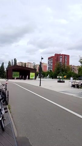 This media file captures a street-level view of an outdoor seating area for a cafe or restaurant, likely in Orekhovo-Zuyevo, Russia, given the location context. The scene is set on a paved walkway adjacent to a road. There are numerous black tables and chairs arranged outdoors, suggesting a public space where people can gather. In the background, there are multi-story buildings, some of which appear to be residential, with distinct architectural styles. Lush green trees are interspersed throughout the scene, adding to the urban park-like atmosphere. The sky is overcast with thick clouds, indicating a cloudy or possibly rainy day. The lighting is diffused, typical of an overcast day, and it's difficult to determine the exact time of day, but it appears to be daytime.

There are no prominent human figures actively engaged in the scene, although a few individuals are visible sitting at tables in the distance, appearing relaxed or engaged in conversation. A notable object is a wooden kiosk or small structure with bright green signage, featuring text that is partially legible. The visible text on the signage includes "Лото" (Loto), which is likely related to a lottery or game. Another sign with similar green lettering is also present. The overall mood of the media file is somewhat subdued due to the cloudy weather and the lack of dynamic activity, presenting a relatively calm urban environment. The presence of outdoor seating and the surrounding buildings suggest a commercial or mixed-use area within the city.