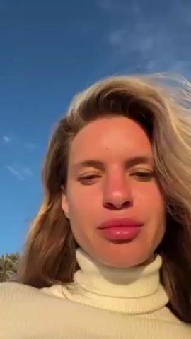 A woman with light skin and blonde hair, wearing a white turtleneck sweater, stands outside on a sunny day. Her face appears slightly sunburned, especially around her nose and cheeks. Behind her, palm trees sway in the breeze against a clear blue sky. The video appears to be a selfie, focusing closely on the woman's face. She looks directly at the camera, occasionally pursing her lips slightly. The background hints at a tropical or coastal location.