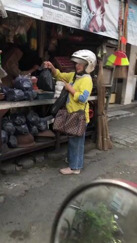 The video shows a person wearing a white helmet, a yellow long-sleeved shirt, and blue jeans standing in front of a market stall. The person is holding two black plastic bags, one in each hand. The market stall is filled with various goods, including bags of what appear to be groceries and other items. A brown and tan checkered shoulder bag is slung across the person's body. The ground is paved with bricks. In the foreground, a red reflective surface, possibly a motorcycle mirror, is visible, showing a reflection of green foliage. A broom and some other items are leaning against the stall in the background. The overall setting appears to be an outdoor market.