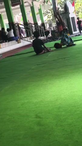 Two children are on a green carpeted surface. One child, wearing a black shirt and grey pants, is kneeling on their knees. The other child, wearing a yellow shirt and a patterned skirt, is in a kneeling position with their hands on the ground and their legs extended behind them, appearing to be in the process of an acrobatic move. There are other people in the background, some sitting and others standing. There is a building with green pillars in the background, and a railing. The environment appears to be outdoors, possibly in a courtyard or open area adjacent to a building.