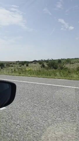 The video shows a view from inside a car driving on a road. The car is in motion and the landscape outside is visible. The side mirror of the car is visible in the foreground, reflecting the interior of the car. A white car is seen driving in front of the car from which the video is being recorded. The landscape outside consists of green fields and hills under a cloudy sky. The interior of the car shows parts of the dashboard and the window frame.
