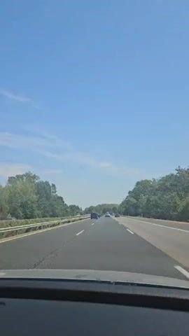 A first-person perspective video shows a clear blue sky with scattered white clouds. The camera is inside a vehicle, looking forward through the windshield onto an empty highway. Several cars are visible driving in the distance on the same road. On either side of the highway, there are dense green trees. The road has multiple lanes and white lane markings. The overall impression is a sunny day while driving on a highway.
