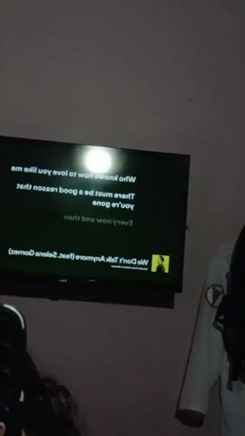 A dimly lit room features a large flat-screen television displaying lyrics to a song. The lyrics are in white text on a black background and are oriented to be read upside down. The song title and artist, "We Don't Talk Anymore (feat. Selena Gomez)," are visible at the bottom of the screen. To the right of the television, a person wearing a white lab coat with patches is partially visible. In the foreground on the left, a person's head and shoulder are visible, with dark hair. The overall lighting suggests it is nighttime or the room is dark.