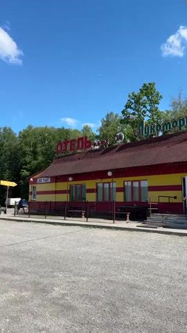 The video shows a bright, sunny day with a clear blue sky and a few white clouds. The camera is focused on a roadside establishment with a red, gabled roof. The building's facade is painted in alternating horizontal stripes of yellow and red. There are several red-framed windows and a prominent red sign on the roof that reads "ОТЕЛЬ" (HOTEL) in large letters, with a smaller sign next to it indicating it is open 24 hours. To the right of the hotel sign, another red sign reads "ЛУКОМОРЬЕ" (LUKOMORYE). A white vending machine dispensing water, labeled "ВОДА" (WATER), is situated on the sidewalk in front of the building. To the far right, a smaller wooden structure appears to be a kiosk or stand with some signage. The ground in front of the establishment is a rough, gray gravel lot. Trees and foliage are visible in the background behind the building. A parked vehicle is partially visible on the left side of the frame. There are no people clearly visible in the video. The overall impression is that of a functional, possibly budget-friendly, roadside stop.