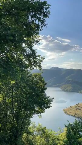 The video captures a panoramic view of a serene lake surrounded by lush green, rolling hills and mountains under a clear blue sky with scattered clouds. The foreground is dominated by the vibrant green foliage of trees and bushes, framing the expansive landscape. The lake itself is calm, reflecting the sky and the surrounding topography. The terrain is varied, with some slopes covered in dense forest and others with sparser vegetation, revealing patches of dry, golden grass. The sunlight casts gentle shadows, highlighting the contours of the land and adding depth to the scene. The overall impression is one of natural beauty and tranquility. No people or man-made objects are clearly visible.
