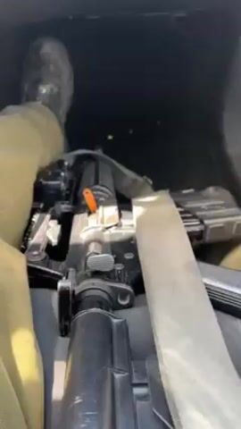 Here's a detailed description of the video:

**Setting:** The video appears to be taken inside a car. The dashboard, steering wheel, and gear shift are visible. The audio suggests music is playing through the car's sound system. 

**People:** Only a person's legs and hand are partially visible in the video. The person is wearing khaki pants and black shoes.

**Objects:** The video shows various objects inside the car:
*   A touchscreen display showing what seems to be a music player app.
*   A set of car keys.
*   A small, white container filled with what looks like dried, possibly crushed, plant material, with a greenish-brown color.

**Actions:**
*   The video starts with a view of the gear shift and the person's leg.
*   The camera angle shifts to show the dashboard display and the music playing on it.
*   The camera then focuses on the container held in the person's hand.

**Environment:** The lighting indicates it's likely daytime, with sunlight visible in the car.