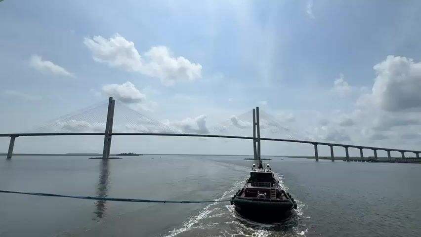 A tugboat is moving on a wide river towards a large cable-stayed bridge. The tugboat is pulling a blue line. The sky is blue with white clouds, and the water is choppy. The bridge has two tall towers and a long span. The tugboat is leaving a white wake behind it. The video is shot from a slightly elevated perspective.