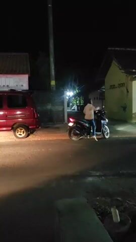 A person on a motorcycle turns on their headlight and begins to ride away from a parked red vehicle. The scene takes place at night on a paved road in front of buildings.