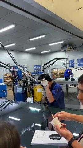 In an industrial setting, a man in a blue uniform is using a tool connected to a complex machinery system involving a blue container and a yellow barrel. He's wearing gloves and has earphones in. He appears to be applying something to a surface in front of him, while a hand holds up a cup and a document is on the surface. The scene is lit with fluorescent lights and includes another person in a blue uniform working in the background.