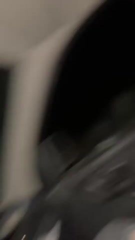 A young woman with dark hair tied back sits in the front passenger seat of a vehicle at night. She is wearing a white t-shirt with colorful text and a dark jacket. Her head is tilted downward, and she appears to be looking at something in her hands, though the object is not clearly visible. Her right hand, with pink nail polish, is seen in the lower right of the frame. The vehicle's door panel and a dark window, reflecting a faint, indistinct shape, are visible. The scene is dimly lit.
