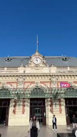 A young woman in a white puffer jacket, black pants, and a striped shirt walks towards the camera, smiling slightly, in front of the Nice-Ville train station in France on a sunny day. Other pedestrians are walking near the station entrance. The station is beige and red brick with a large clock on top, displaying the time. A French flag flies above the clock. An SNCF logo is visible. The sky is clear and blue.