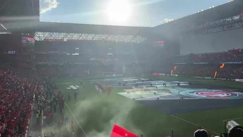 A soccer game is about to begin, with the stadium packed with fans mostly wearing red. Smoke drifts across the field, and a large banner is displayed in the stands that reads "Cree Siempre Rojo". The sun is bright, causing lens flare. Two teams are lined up on the field, facing each other. One team is wearing red and white uniforms. The other team is wearing yellow and blue uniforms. White objects are placed on the field between the two teams, presumably coffins.