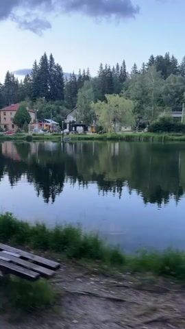The video depicts a tranquil pond at dusk. The water is calm and reflects the surrounding trees and buildings. A weathered wooden bench sits on the edge of the pond, in the foreground.  The sky is overcast, and the light is soft. There are no people visible in the frame. The camera movement suggests the videographer is panning to their right, capturing the scenery. The overall impression is one of peacefulness and quiet.