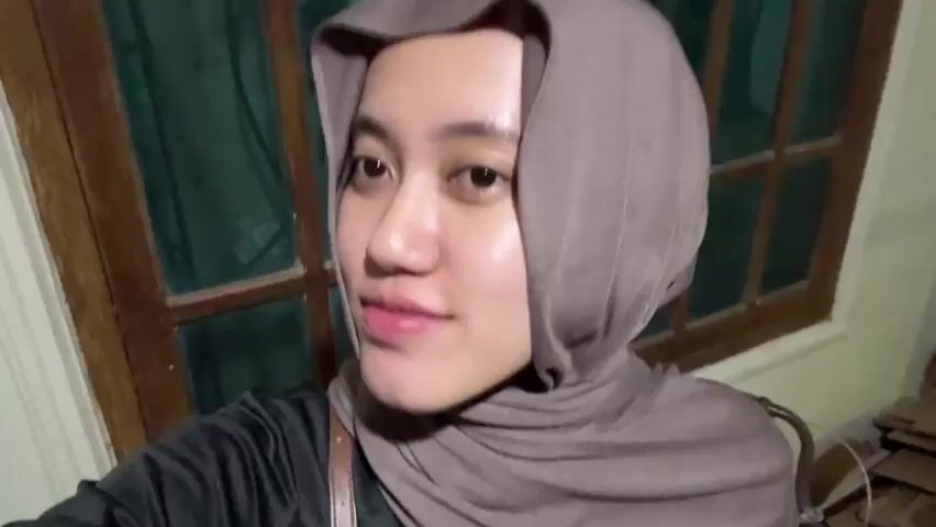 The video shows a young woman wearing a hijab and a black shirt, smiling and looking at the camera. She has braces on her teeth. The background appears to be indoors, with a window frame visible. The lighting is dim.