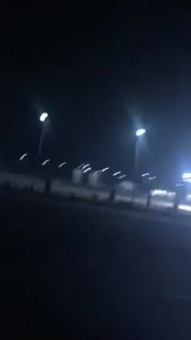 The video depicts a night scene at a gas station or rest stop. The area appears mostly deserted, lit by overhead streetlights and the bright lights of the gas station canopy. The camera is stationary, possibly recording from within a vehicle, and the scene is somewhat dark with the light sources causing bright glare. There is a sense of stillness and isolation.