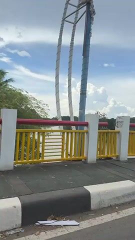 A video taken from a moving motorcycle on a bridge shows the railing of the bridge and the surrounding landscape under a cloudy sky. The railing is white and yellow with red accents on top. Trees and a body of water are visible on the other side of the railing. There is a motorcycle with a trailer in front of the camera.