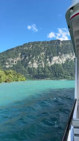 The video shows a view from a boat moving on a bright blue body of water. On the left side of the screen, a steep, tree-covered mountain rises dramatically. The water is a vibrant turquoise color, with small waves and ripples visible. In the distance, along the shoreline, there are patches of green trees and some buildings. The sky is a clear blue with a few white clouds. The right side of the frame shows the railing and part of the boat the video is being filmed from. The camera pans across the scenery, offering glimpses of the shoreline with more trees and distant mountains.