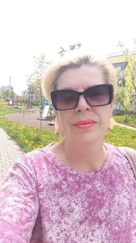 A blonde woman wearing sunglasses and a pink patterned t-shirt is filming a selfie video outdoors. She is walking on a paved path surrounded by grass, bushes, and dandelions. In the background, there are apartment buildings and a playground.
