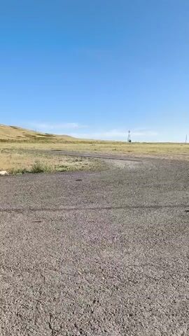 A wide shot captures a desolate, unpaved area with dry grass and a dirt road. In the distance, a lone tower stands against a clear blue sky. The landscape is barren, with rolling hills in the background. The camera pans slowly across the scene, revealing the vastness and emptiness of the environment. There are no people or animals visible in the video, adding to the sense of isolation. The overall impression is one of stark beauty and untouched wilderness.