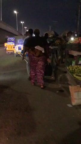 The video shows a night scene, likely a street market or busy roadside. In the foreground, three people are walking away from the camera. The person in the middle is carrying a child on their back. The person on the left is wearing a blue shirt and carrying a rolled-up mat. The person on the right is wearing a dark shirt and a long, patterned skirt. There are market stalls visible on the right side of the frame, with produce like fruits and vegetables displayed. The lighting is dim, with some artificial lights from the stalls and streetlights illuminating the scene. The ground is paved. In the background, there are more people and structures, suggesting a populated area. The overall atmosphere appears to be that of an active, late-night marketplace.
