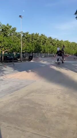 The video captures an outdoor skate park scene featuring a young boy and an adult male skateboarding. The young boy, wearing a white helmet, sunglasses, a dark t-shirt, and light shorts, is seen riding his skateboard across concrete surfaces, standing and balancing on it. An adult male, dressed in a black tank top and light-colored baggy jeans, also skates, moving near a metal rail.

The skate park environment consists of various concrete ramps, ledges, and metal rails. In the background, a line of lush green trees provides a natural border, with parked cars and a street visible beyond. A tall lamppost stands out against the sky, and briefly at the start, the moon is visible in the daytime sky. The sky is clear with a few scattered white clouds, suggesting a pleasant late afternoon or early evening. Graffiti spelling "LICIANS" is visible on a concrete barrier within the park. The overall mood is relaxed, showing individuals engaged in skateboarding activities.